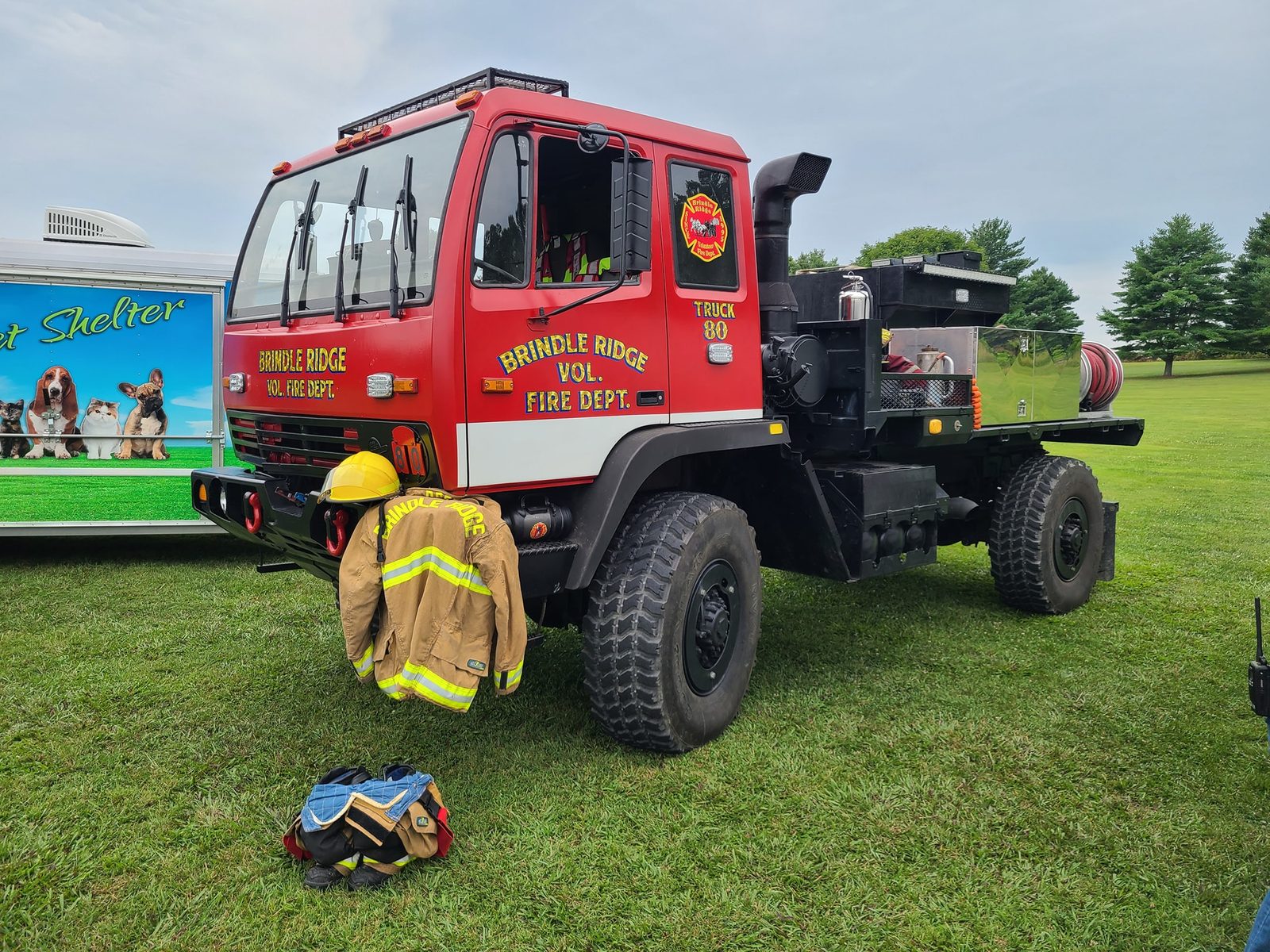 Brush Truck 80, a red military-style 4x4 flatbed brush truck parked on grass with turnout gear hanging from the front bumper