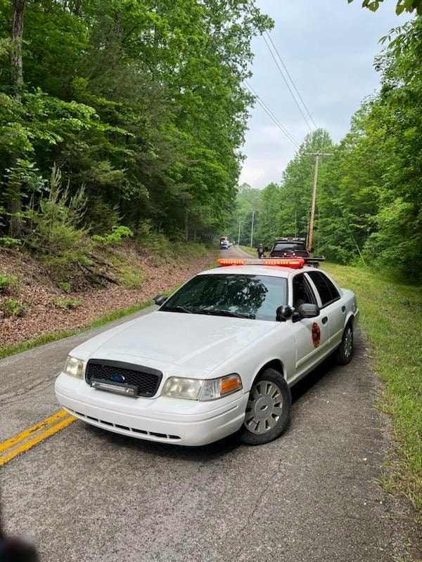 Car 85, a white Ford Crown Victoria with a red lightbar, blocking a narrow tree-lined road at a scene